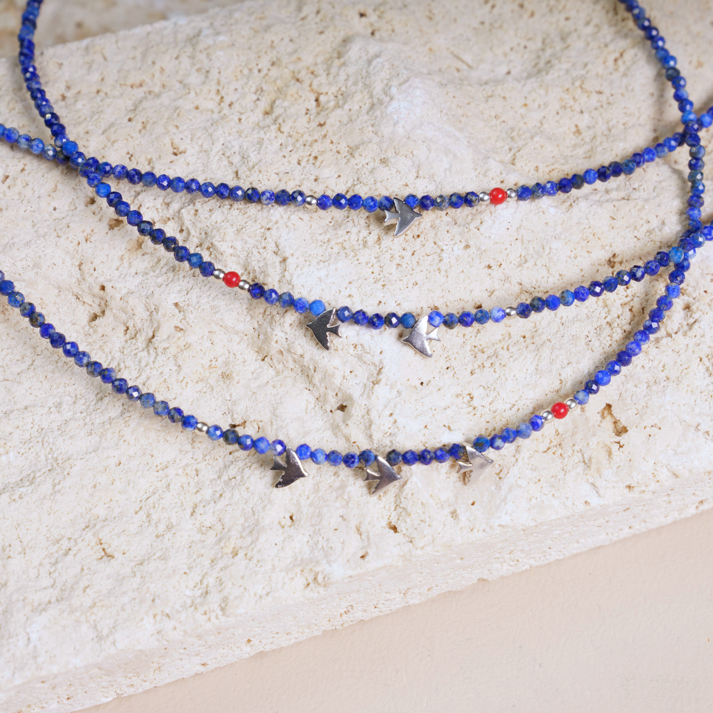 Minimalist lapis lazuli gemstone necklace featuring silver bird-shaped pendants set gracefully on a beige textured surface.