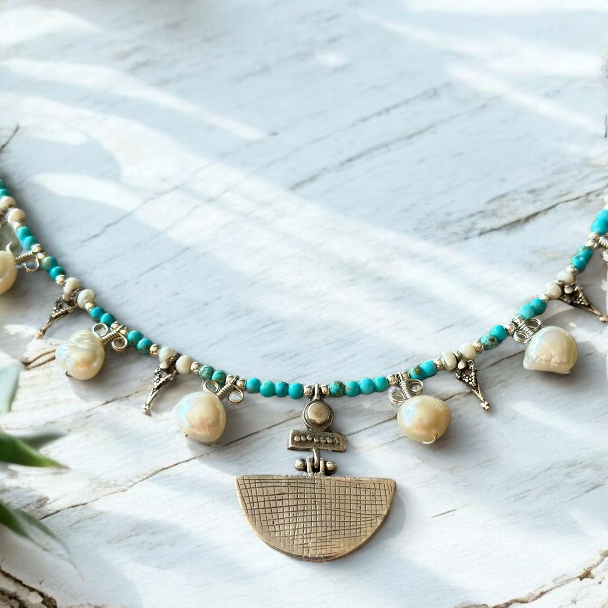 Selene Turquoise Gemstone and Pearl Necklace with intricate metallic pendant on a rustic white surface