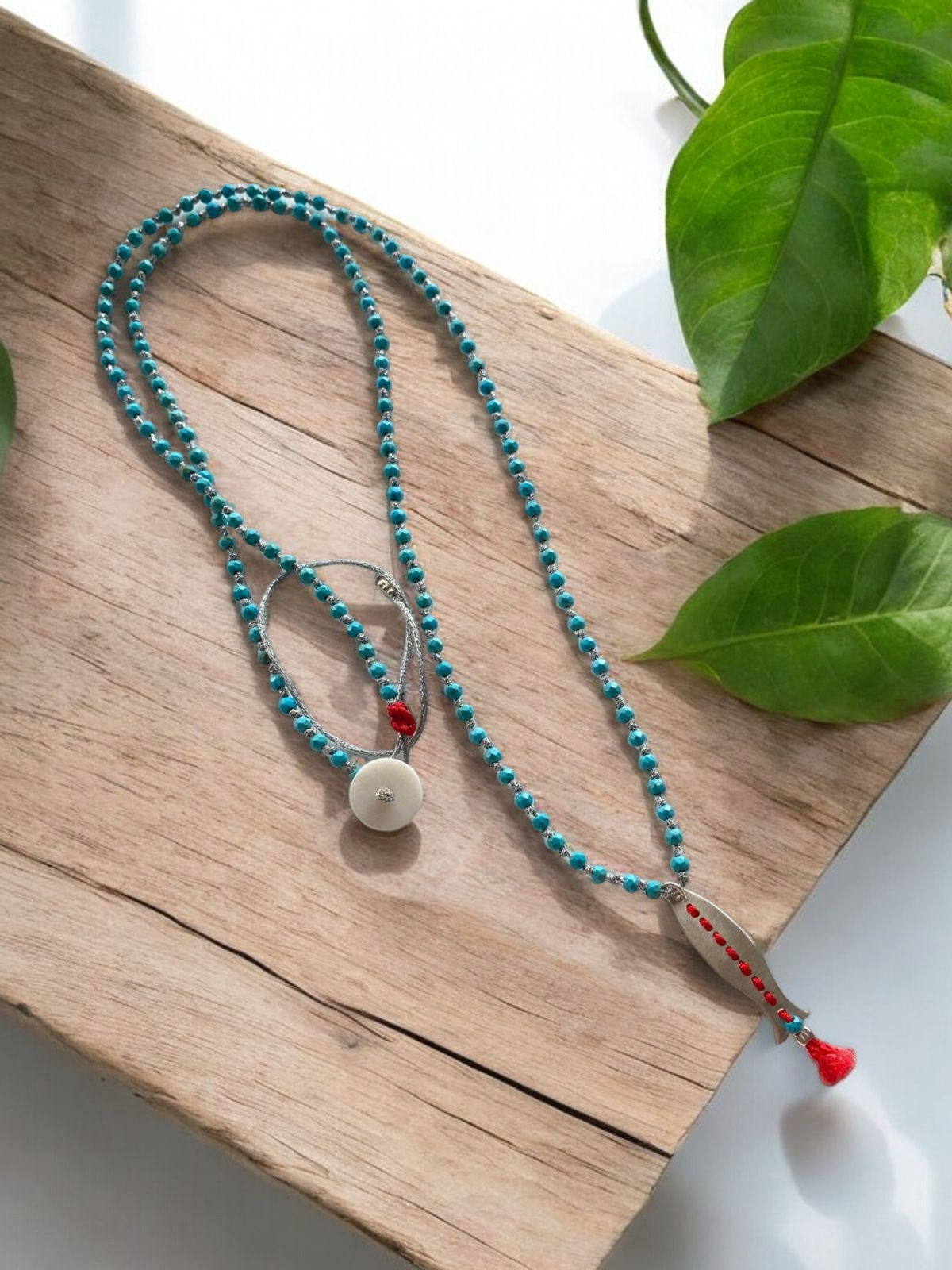 Afrodit Fish Pendant Long Necklace in 925 Silver with turquoise beads, displayed on a wooden surface with green leaves.