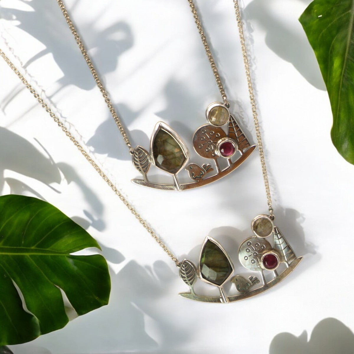 Elegant silver necklaces featuring a nature-inspired design with gemstones, photographed amidst lush green leaves.