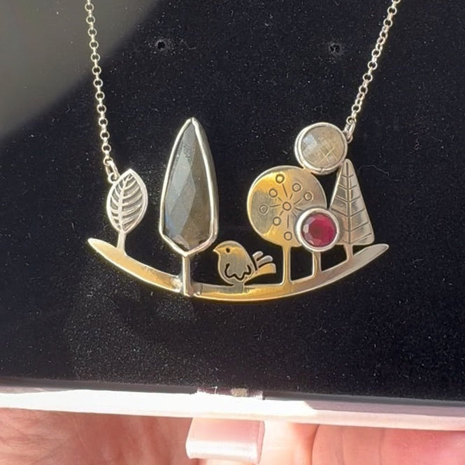 Detailed landscape-themed silver necklace featuring enamelled trees, a bird, and gemstones, presented in a display case.