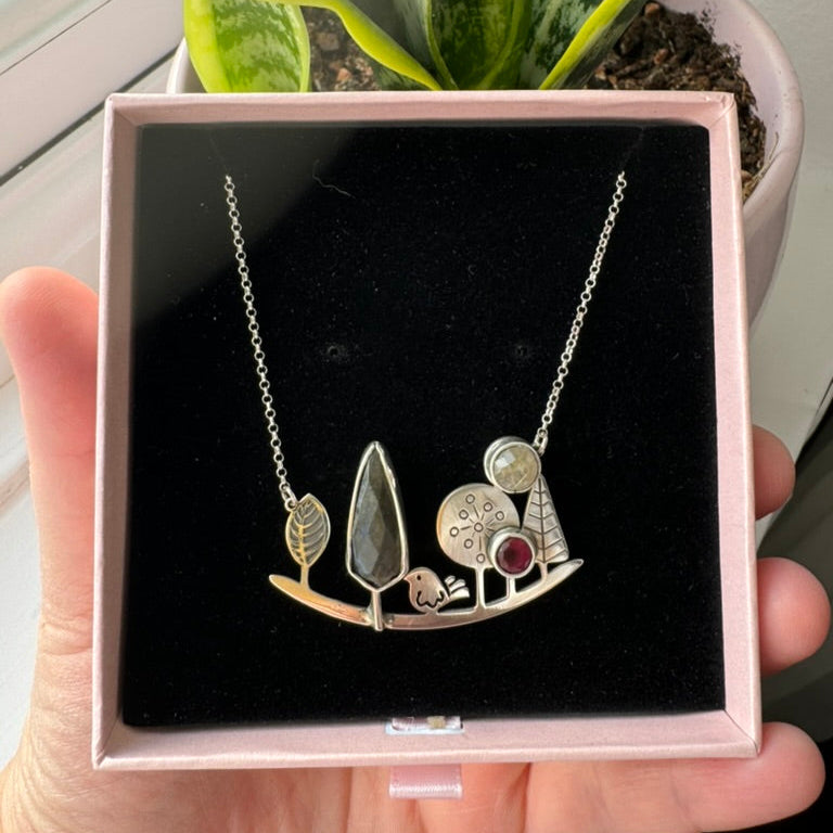 Landscape-inspired silver necklace featuring intricate nature motifs, presented in a jewellery box with a plant in the background.