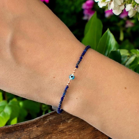 Elegant Lapis Lazuli gemstone bracelet with a mini evil eye charm, showcased outdoors with vibrant greenery and blooms.