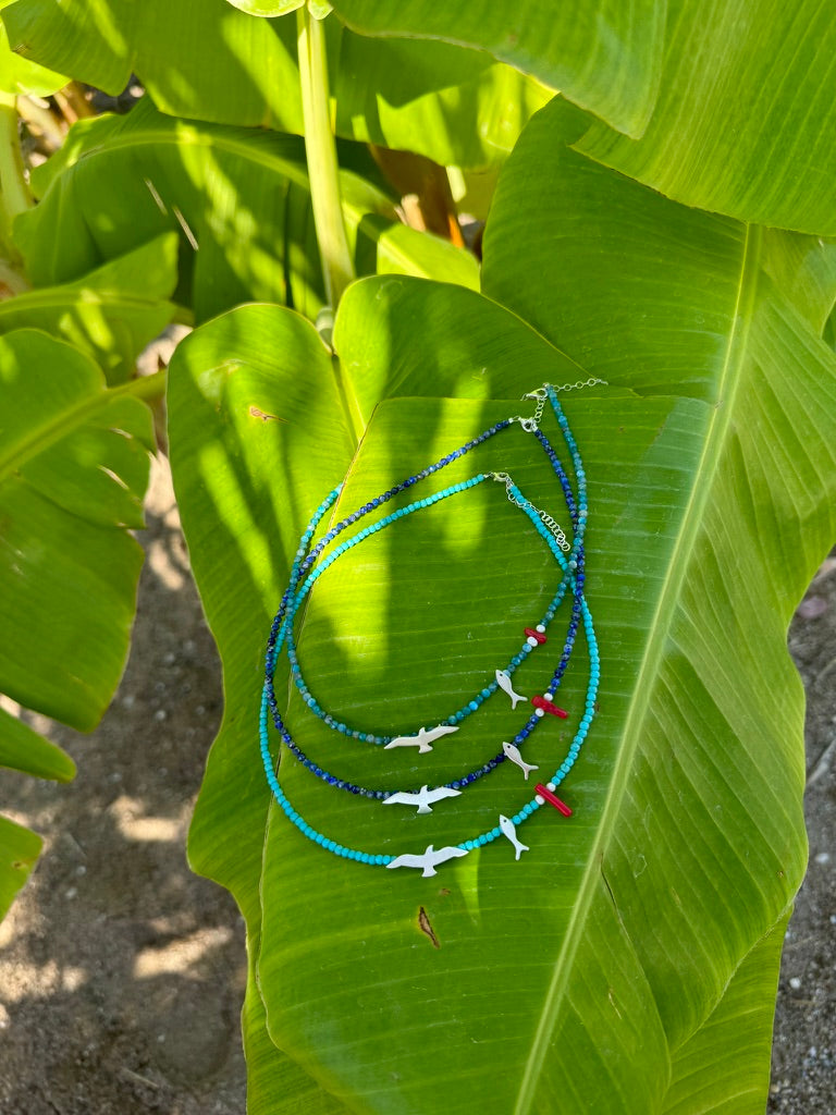 Seagull & Fish Lapis and Silver Necklace displayed on a green tropical leaf under sunlight.