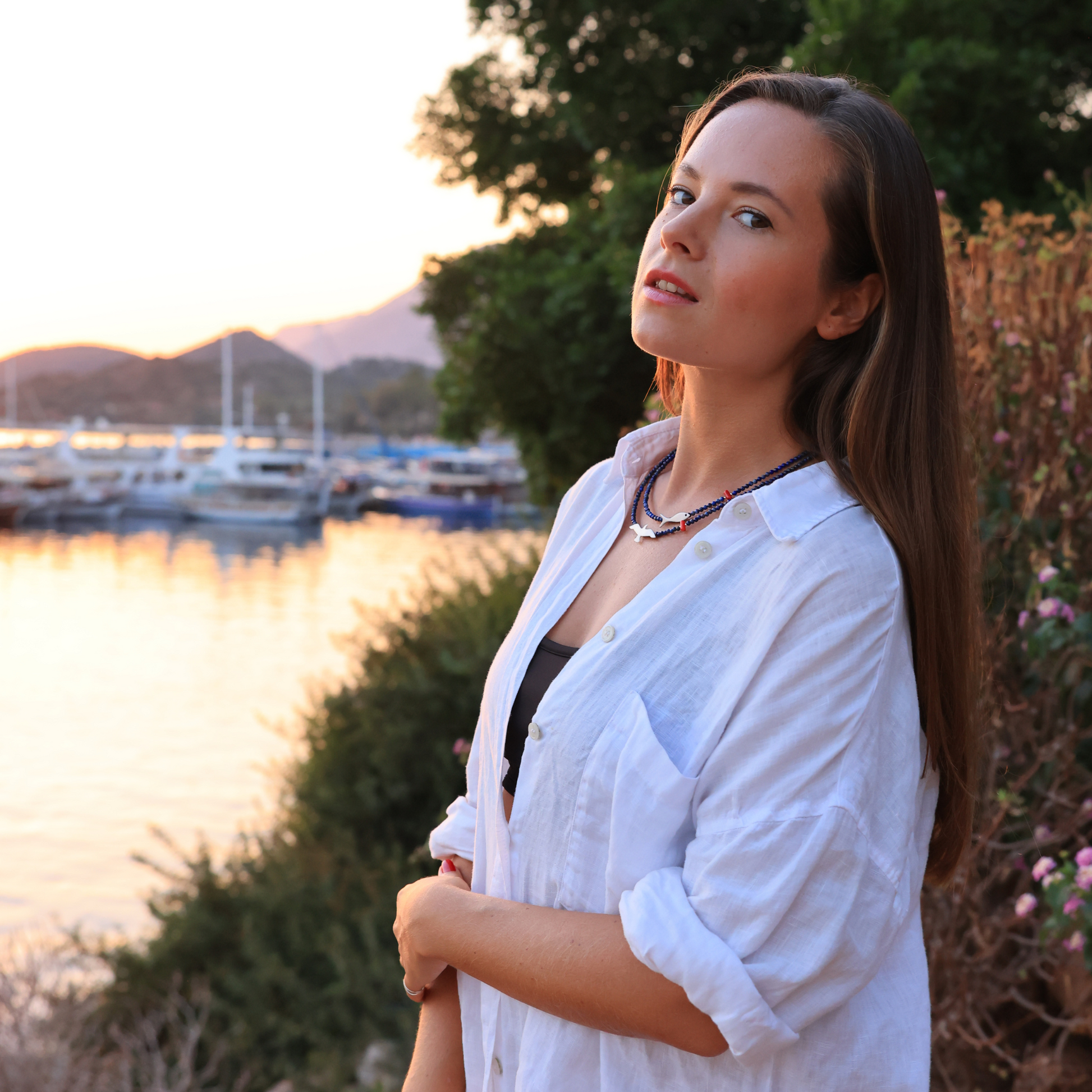Woman wearing a Seagull & Fish Lapis and Silver Necklace, standing by a marina at sunset with a serene background.