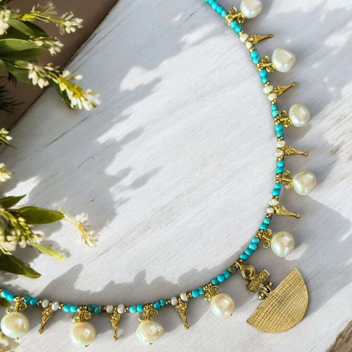Selene Turquoise Gemstone and Pearl Necklace - Gold Plated with intricate design, laying on a wooden surface with flowers nearby.