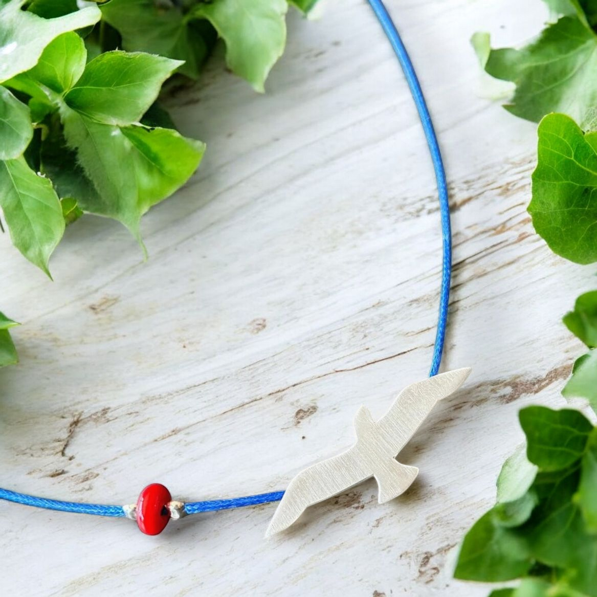 Elegant Silver Seagull Necklace on Blue Cord, Displayed on Wooden Surface with Greenery Accents