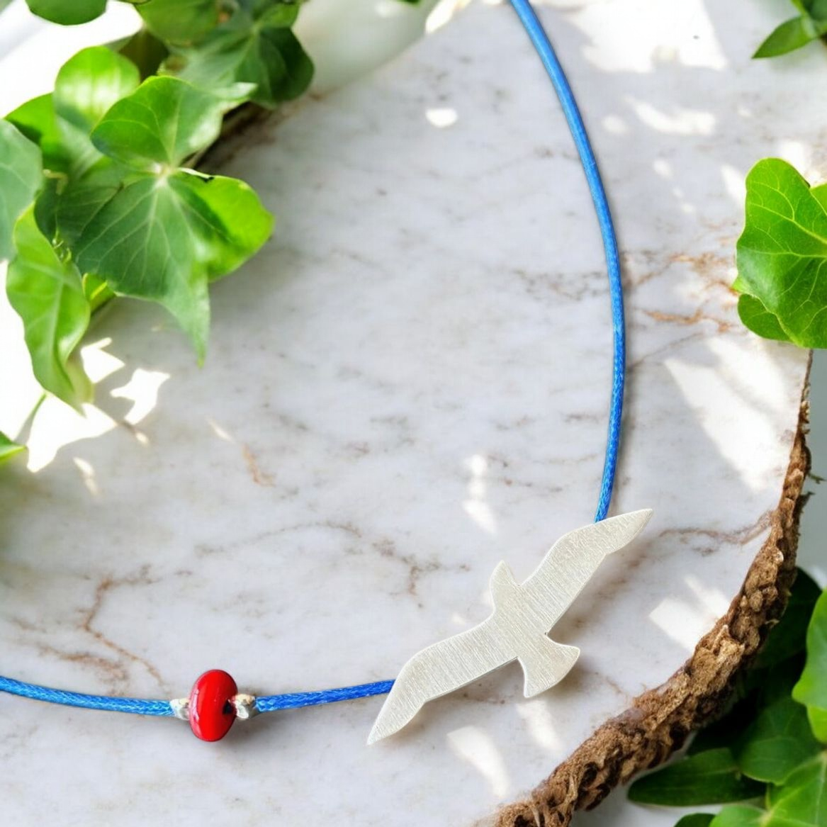 Elegant Silver Seagull Blue Cord Necklace on Marble Background with Greenery for a Nature-Inspired Look.