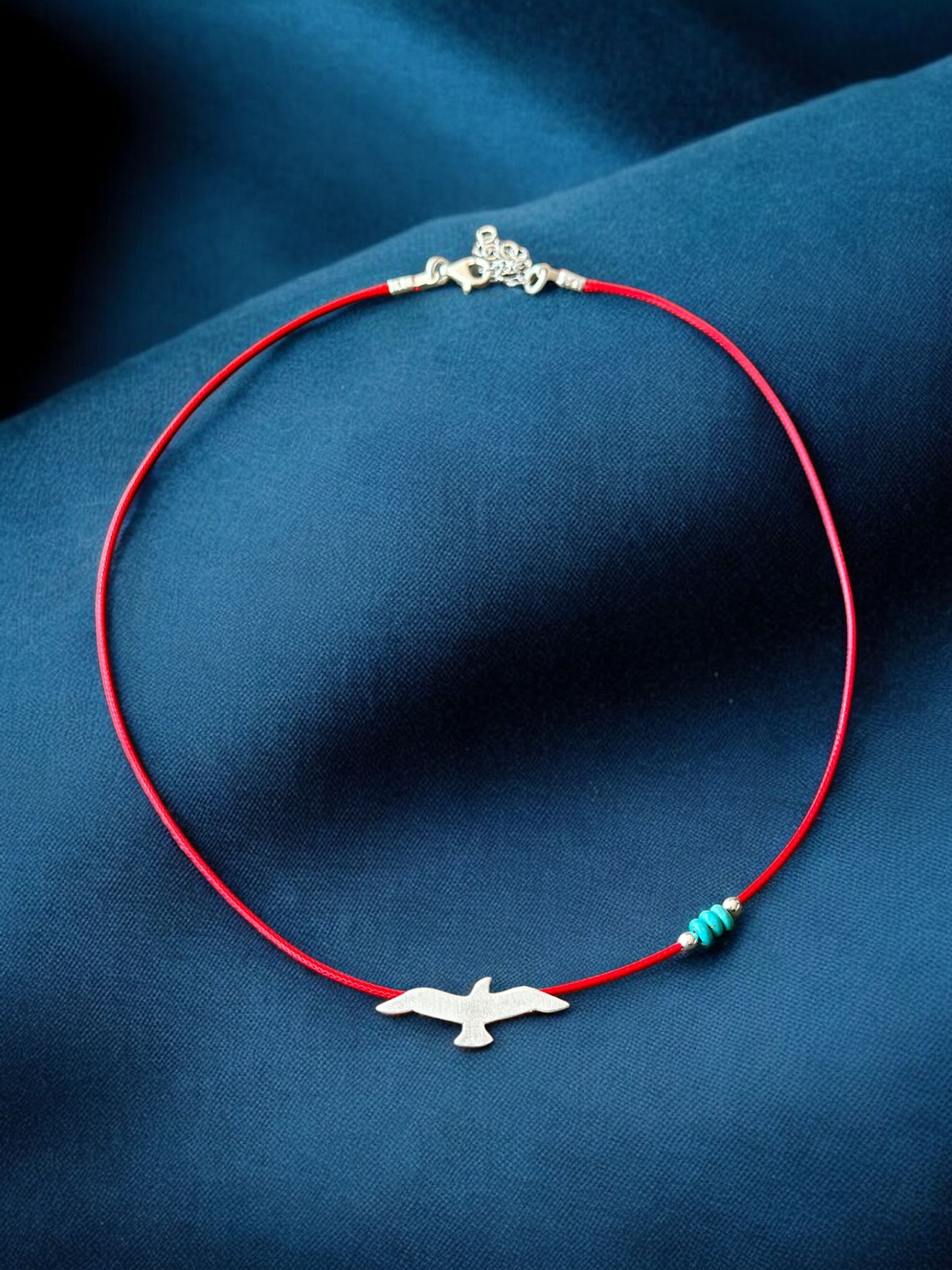 Silver seagull charm on a red cord necklace with gemstone accents, displayed on a dark blue background.