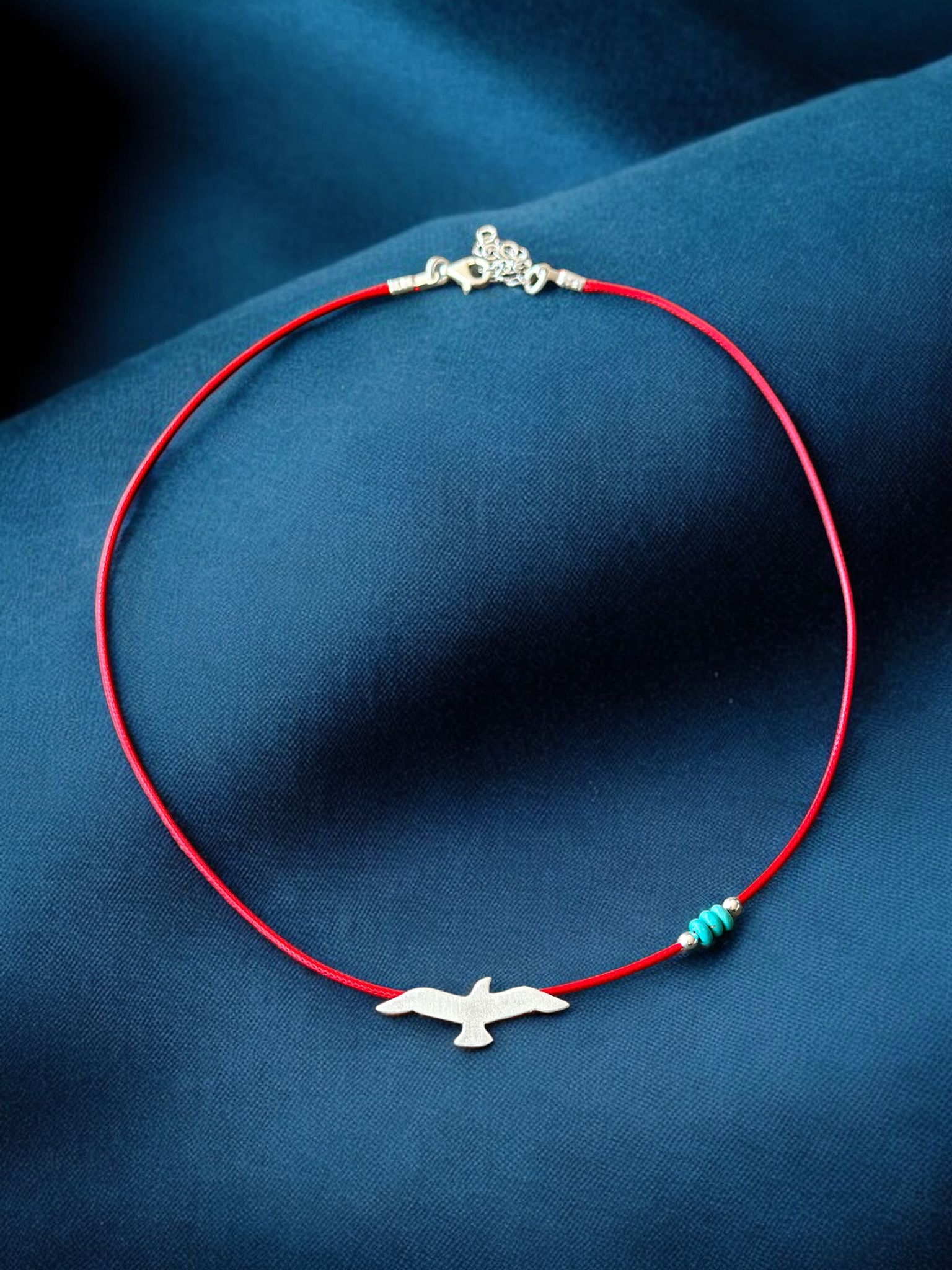 Silver seagull charm on a red cord necklace with gemstone accents, displayed on a dark blue background.