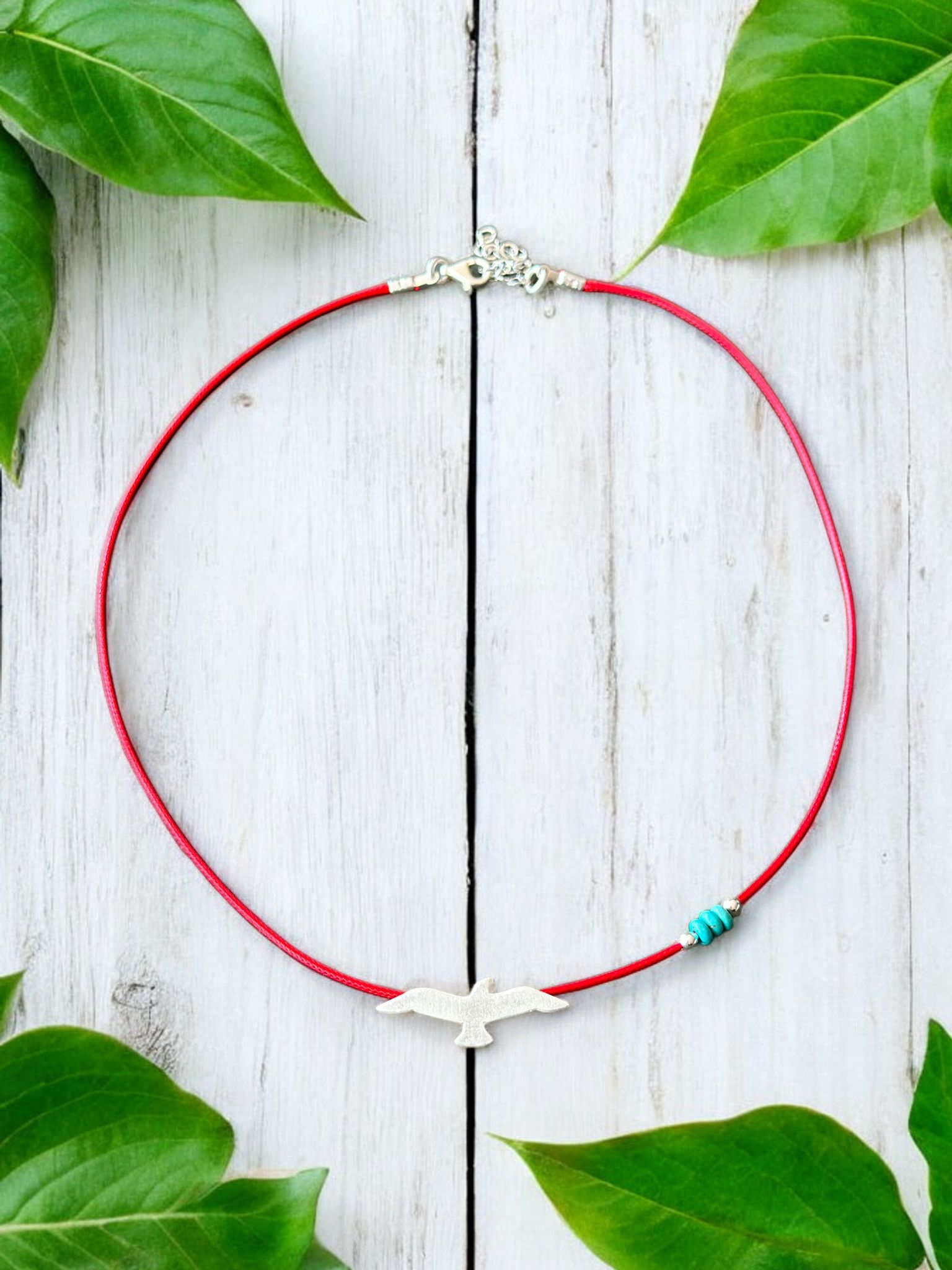 Silver seagull pendant on red cord necklace, elegantly displayed with green leaves on wooden background.